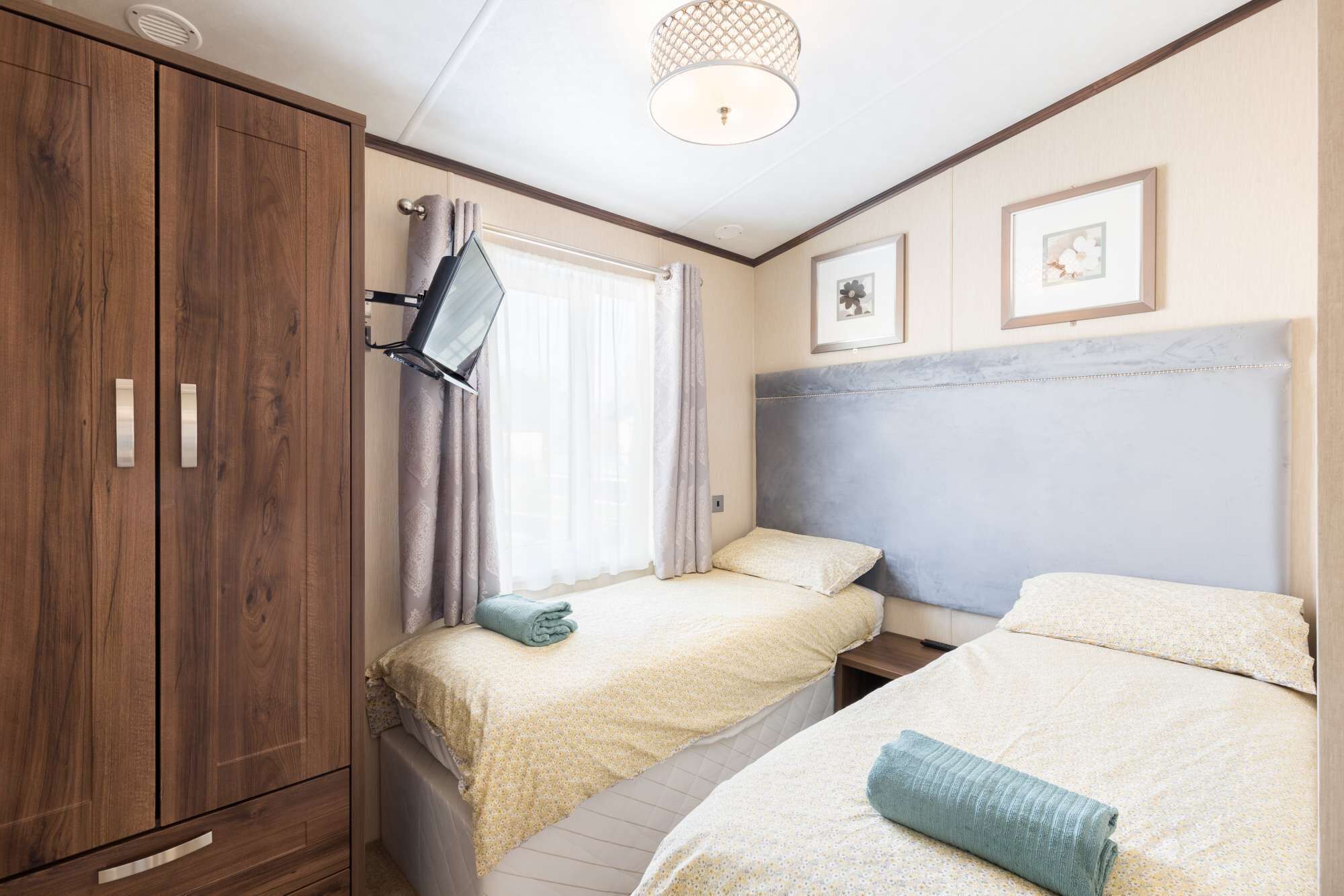 Une chambre avec deux lits, une armoire en bois, un luminaire au plafond et des tableaux encadrés accrochés au mur.