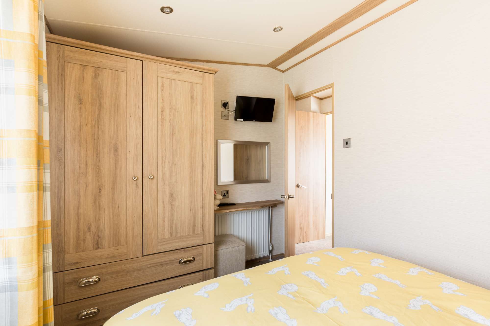 A bedroom with a wooden wardrobe, a bedside table, a mirror, and a bed with a patterned bedspread.