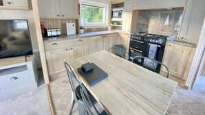 A compact kitchen with wooden cabinets, a built-in oven, and a dining area with a table and chairs. - Burford Hot Tub Holiday Home (Hoburne Holiday Park)