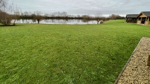 A grassy area with a pond, a wooden building, and leafless trees. - Lilly Lodge on the edge of Lake Pochard (Lake Pochard)