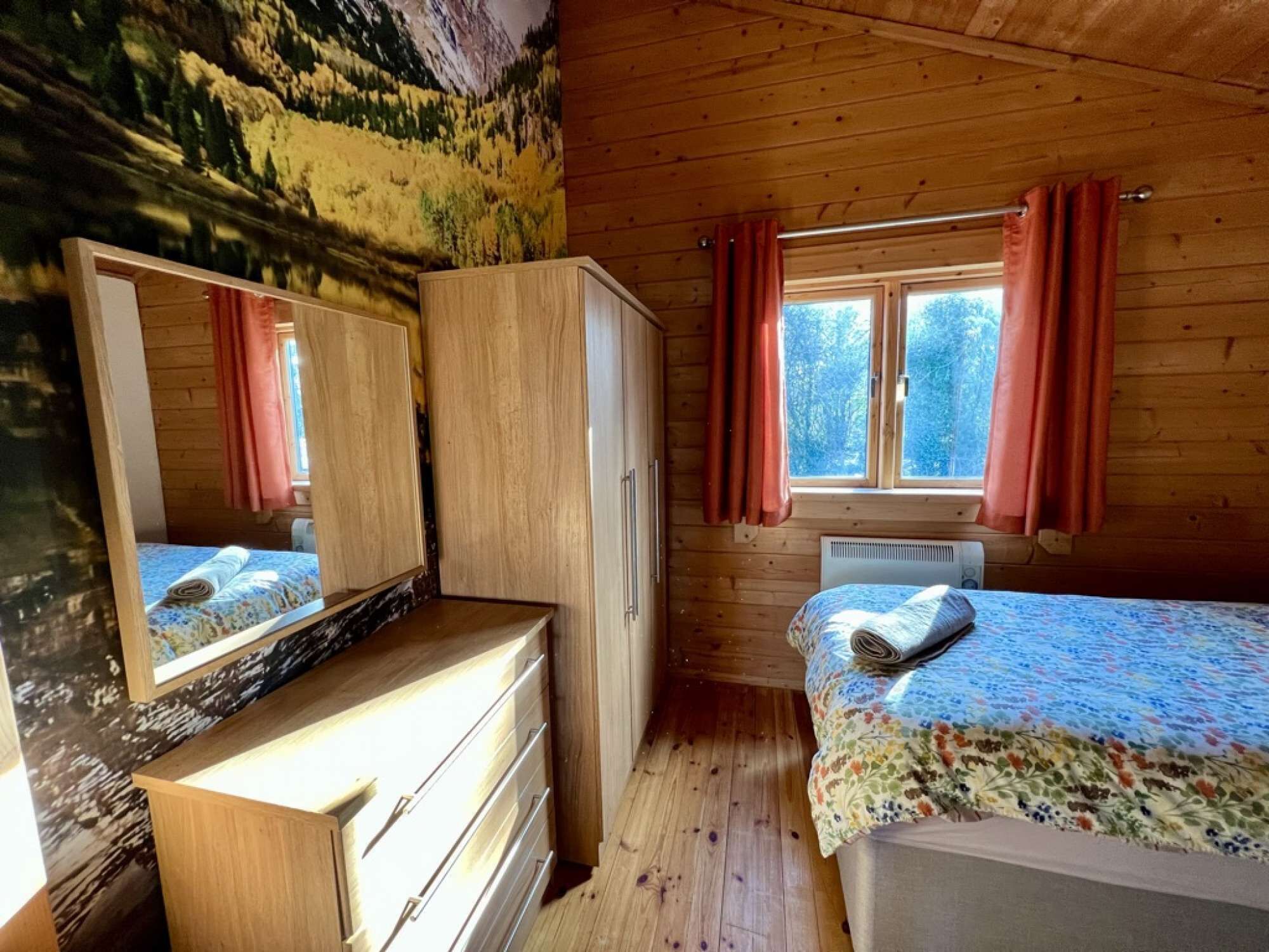 A cabin with a wooden interior, a bed with a floral bedspread, a dresser, a mirror, and a window with curtains.