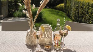 A table set with drinks, a bottle, and decorative items on a balcony with a view of a garden and a building. - Neu Eingerichtetes Apartment mit Pool, Fitnessstudio und Sauna, Nähe Bardolino (Cavaion Veronese)