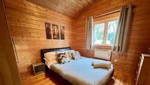 Un dormitorio de cabaña de madera con cama, almohadas, mesita de noche y ventana con cortinas.