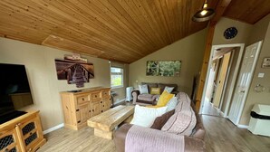 A cozy living room with a wooden ceiling, a sofa with cushions, a wooden cabinet, and a television. - Bumble Bee Cabin on Heron Lake (Hoburne Holiday Park)