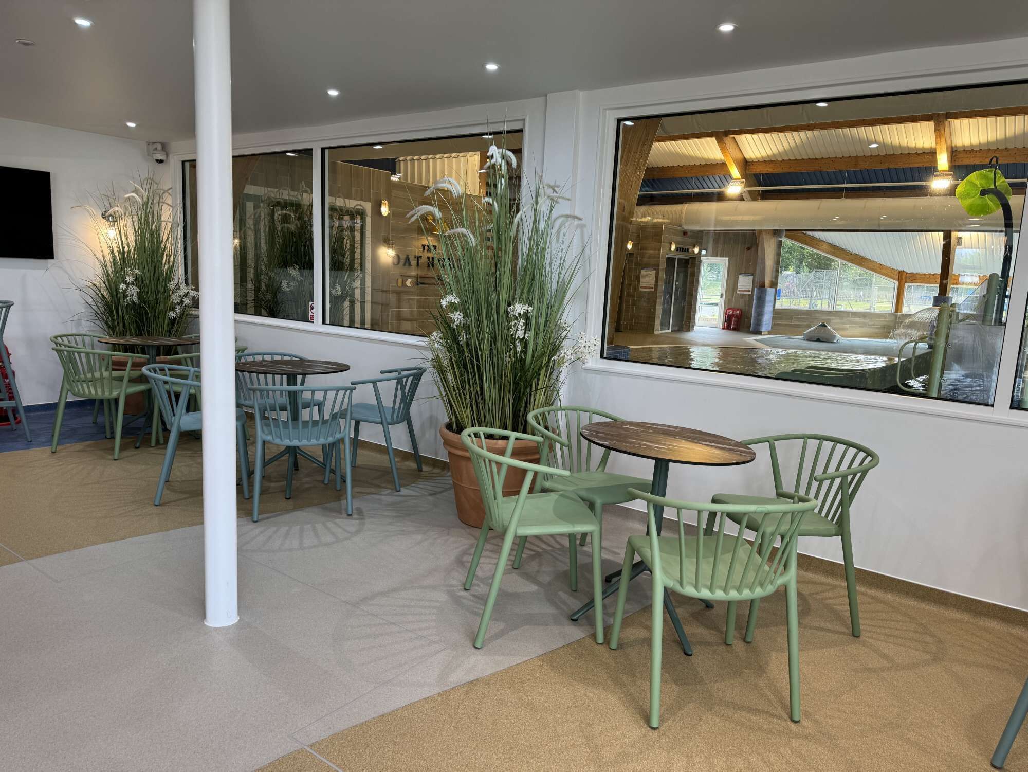 An indoor seating area with green chairs and tables, large windows, and indoor plants.