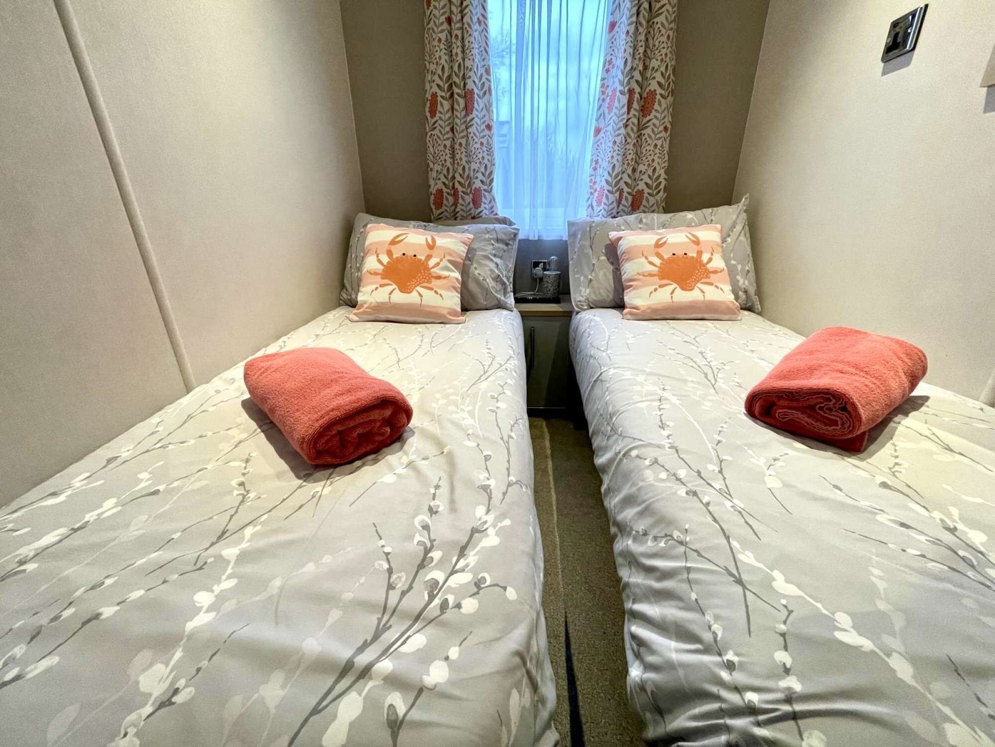 A small, compact room with two single beds, each with a rolled red towel. The beds have floral-patterned bedding. There is a small table between the beds with a cup and a small item on it. The room has a window with curtains, and the walls are a light color.