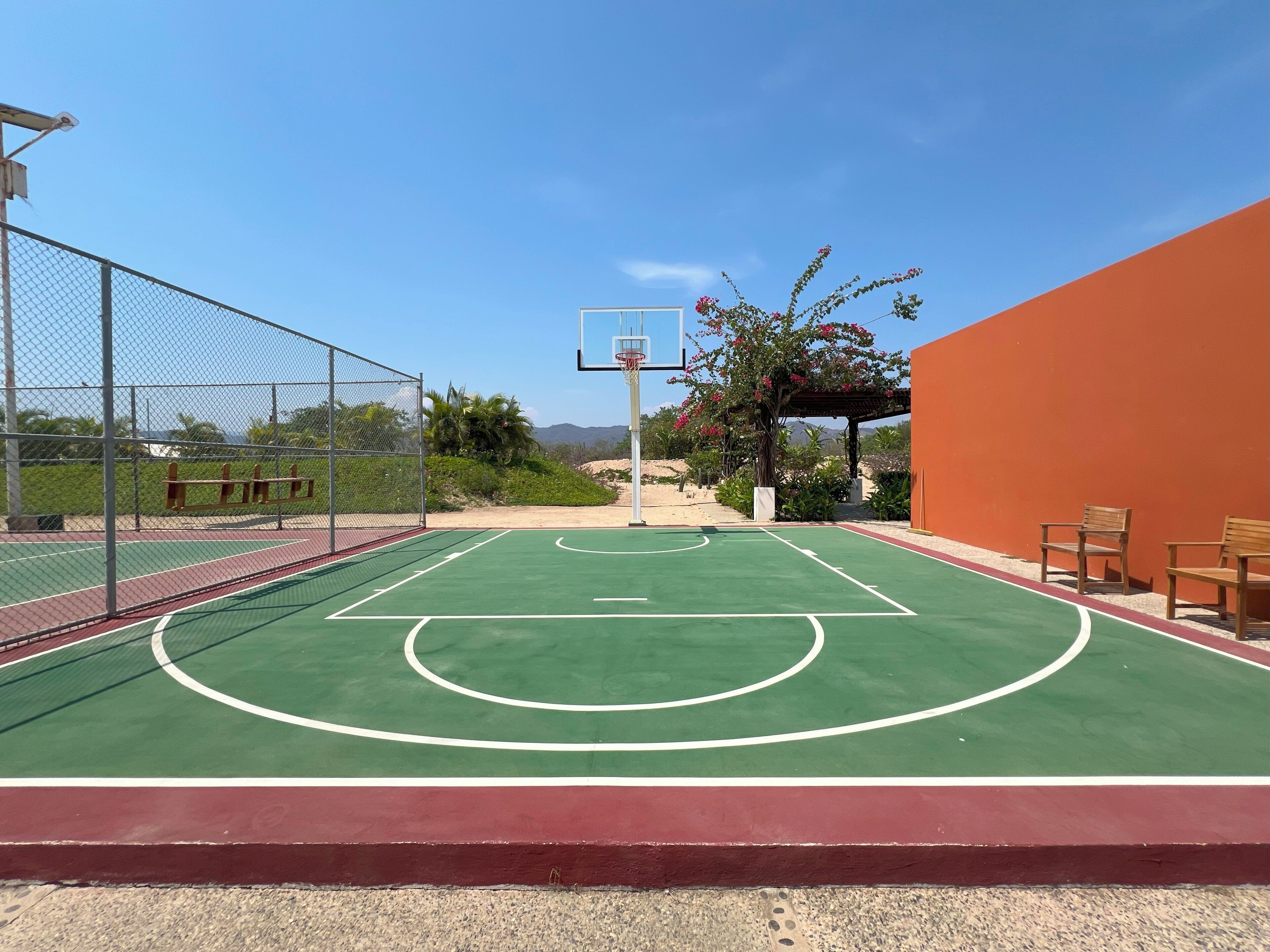 Een basketbalveld buiten, met een basketbalring, omgeven door een hek en een rode muur.