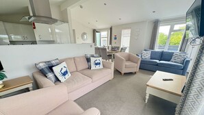 Un salon moderne avec un canapé beige, un fauteuil bleu et un divan bleu. On y trouve une petite table sur laquelle est posée une télécommande. La pièce est dotée d’une grande fenêtre avec des rideaux et offre une vue sur la verdure à l’extérieur.