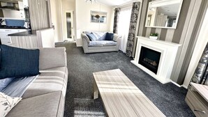 Un salon moderne avec un canapé, une table basse, une cheminée et un coin cuisine visible à travers une porte ouverte.