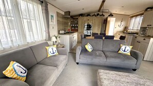 A modern living room with grey sofas and patterned ottoman, a kitchen with wooden cabinets, and a dining area with a round table.