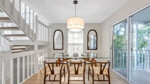A dining area with a glass table and wooden chairs, a staircase with white railings, and large windows with a view of greenery.