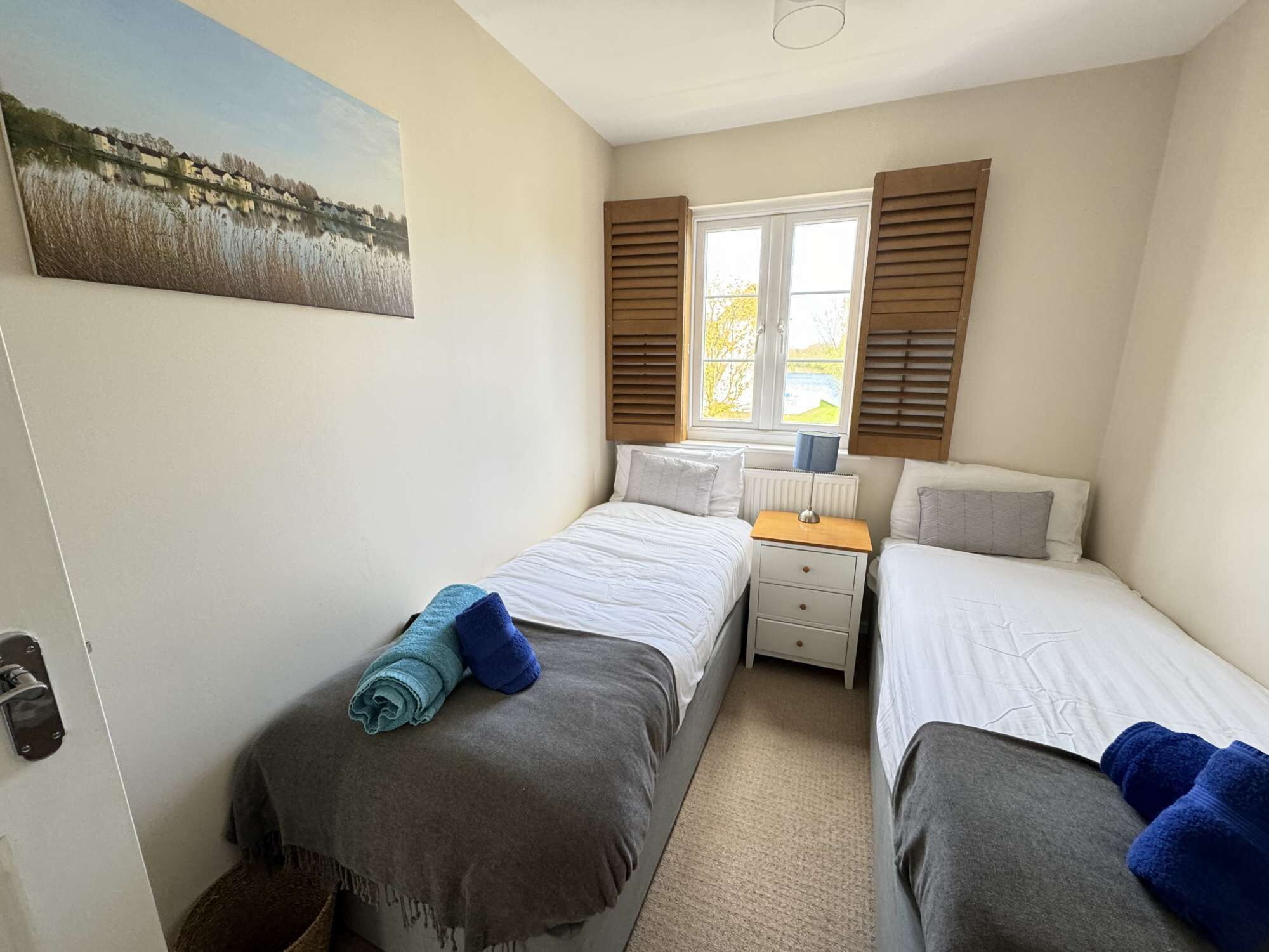 Une chambre avec deux lits, une fenêtre avec des volets, une table de chevet et un tableau au mur.