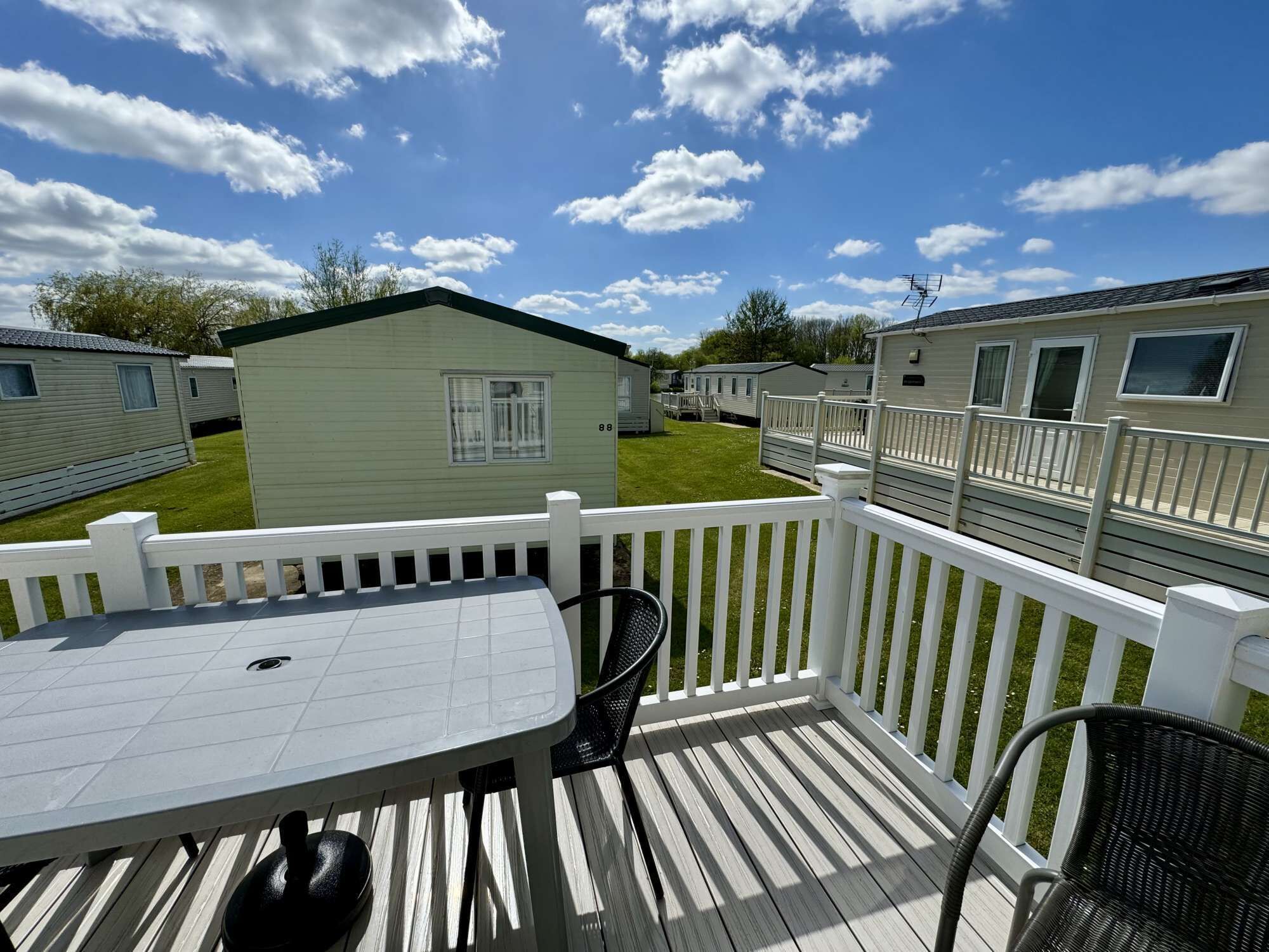 A deck with a table and chairs, surrounded by white railings, overlooking a grassy area with mobile homes.