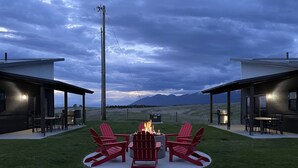 A modern outdoor seating area with a fire pit and red Adirondack chairs, surrounded by a well-maintained lawn and overlooking a mountainous landscape. - The Jewel View Cottages Unit 3, 1BD1BA (Bigfork)