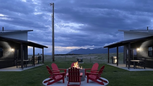 Ein moderner Außenbereich mit Sitzgelegenheit, einem Feuerkorb und roten Adirondack-Stühlen, umgeben von einer gepflegten Rasenfläche mit Blick auf eine gebirgige Landschaft.