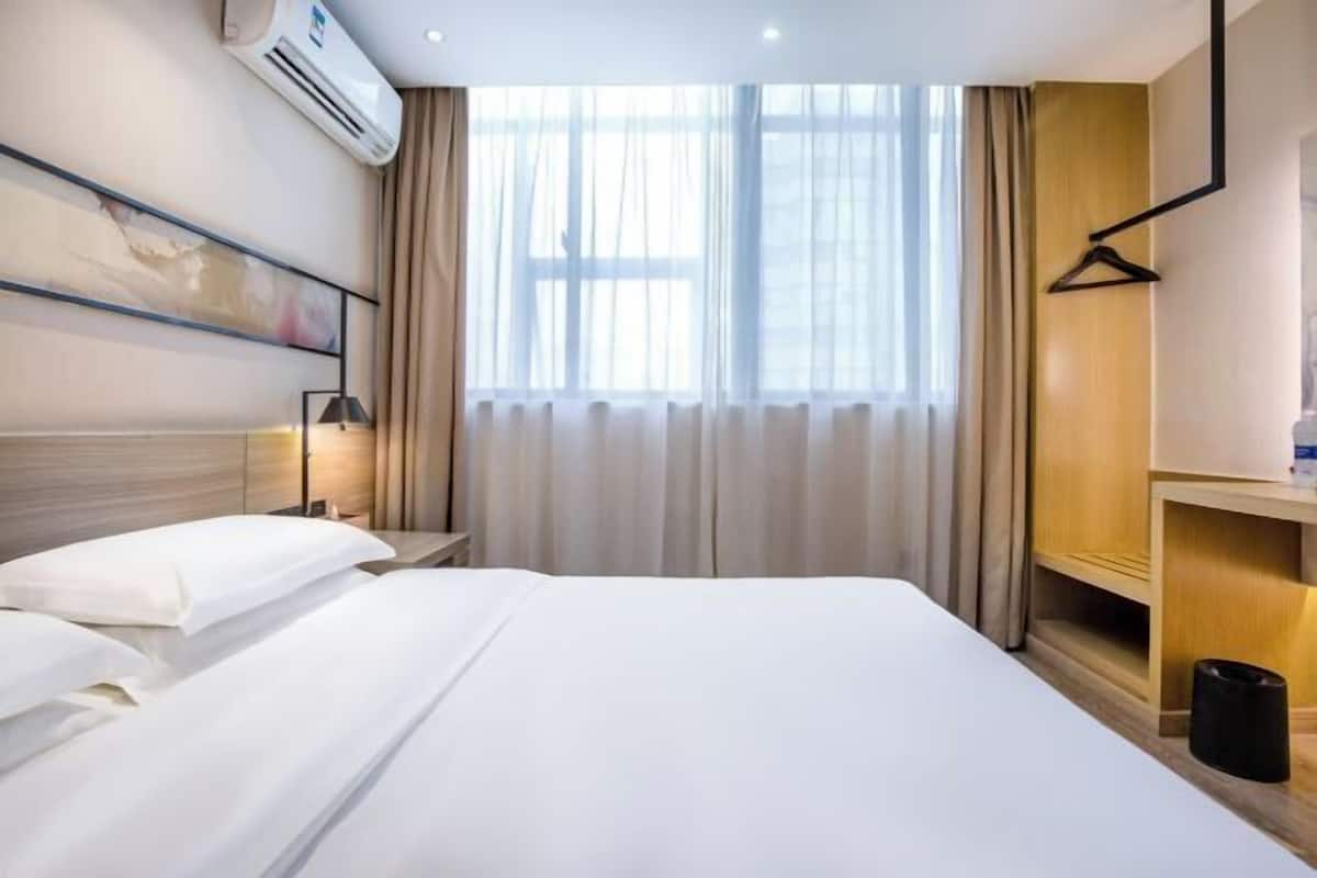 A hotel room with a bed, a bedside table, a lamp, a hanging coat rack, and a window with curtains.