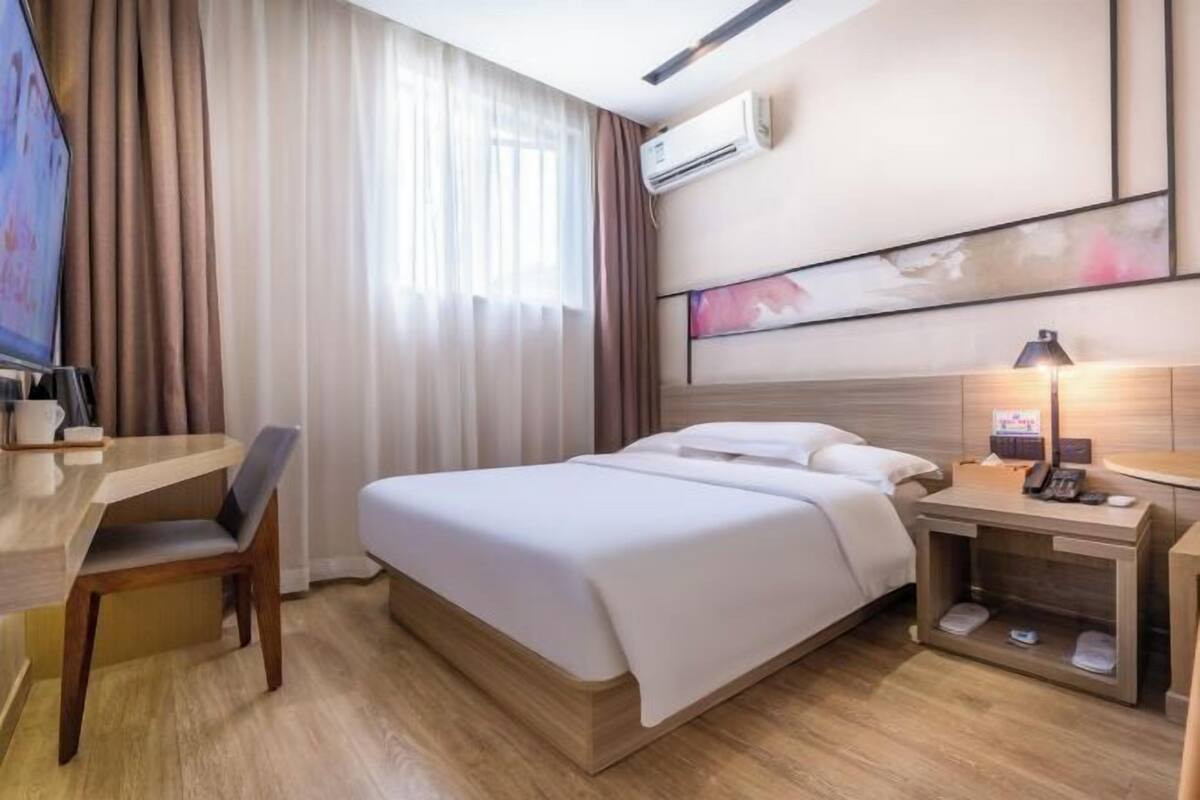 A hotel room with a bed, a desk, a chair, a bedside table with a lamp, and an air conditioning unit on the wall.