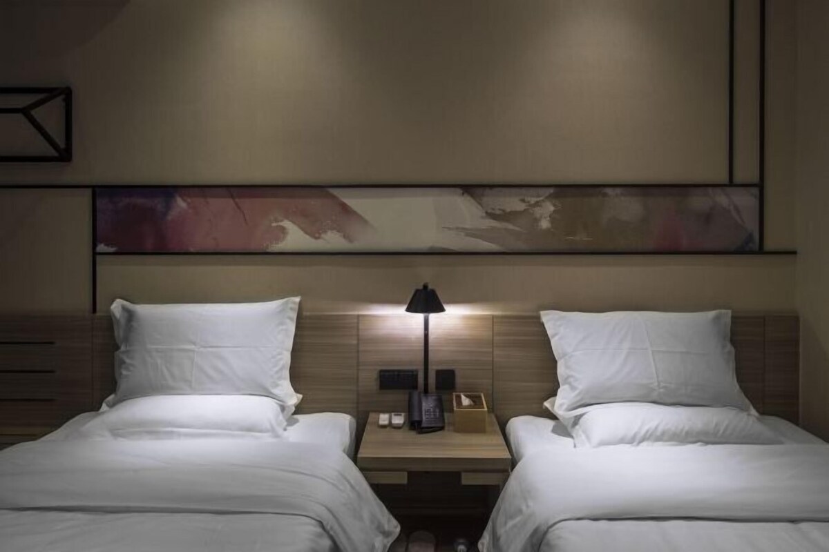 A hotel room with two beds, a bedside table with a lamp, and a wall-mounted artwork.