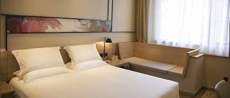Una habitación de hotel con una cama grande, una mesita de noche, un sillón y un ventanal con cortinas.