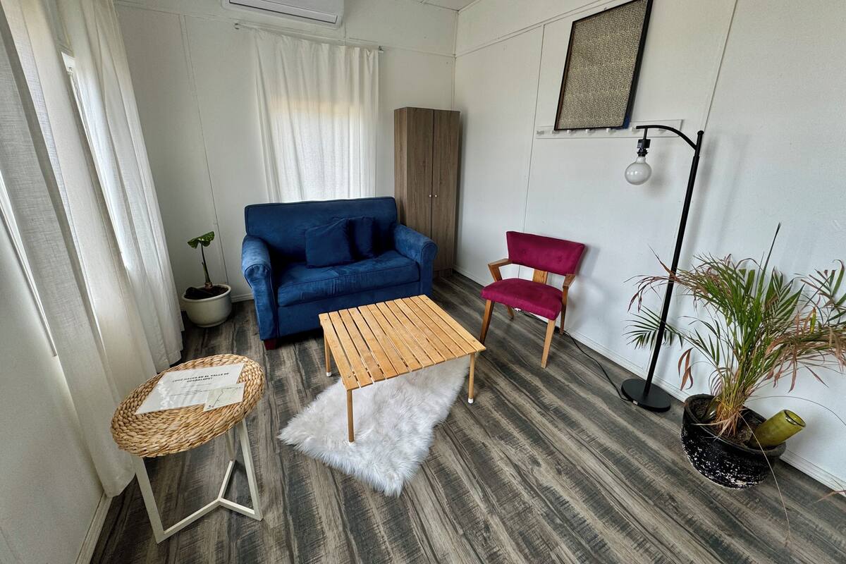 A small, modern living room with a blue sofa, a wooden coffee table, a red chair, and a small side table with a basket. There is a potted plant on the floor and another on a side table.