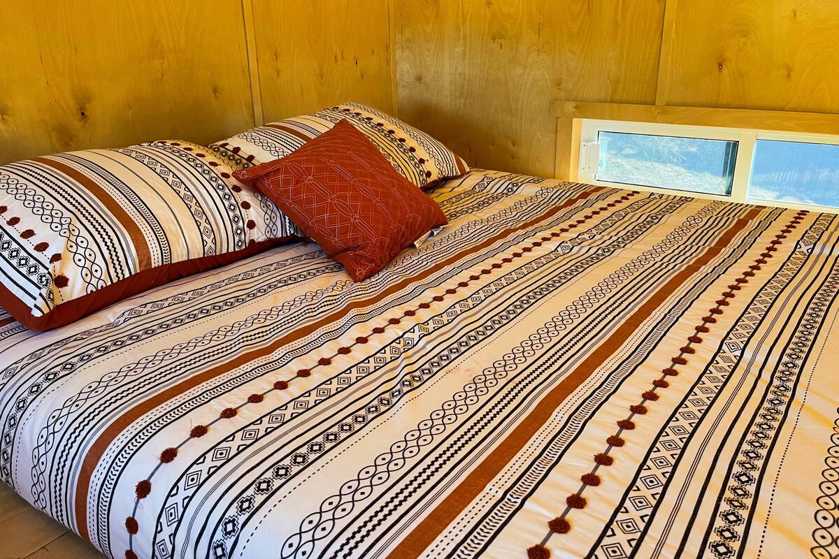 A bed with patterned bedding and a red pillow in a room with wooden walls and a window.