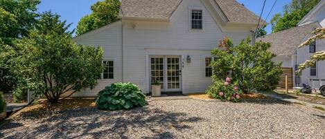 A white two-story house with a gravel driveway, a large green bush, and a flowering shrub.
