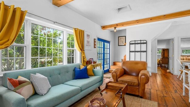 Un salon comprenant un canapé, une table basse en bois et un fauteuil en cuir. De grandes fenêtres sont présentes, ainsi que des rideaux, une porte bleue et un escalier menant à une autre pièce.