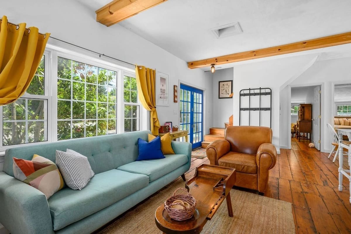 Un salon comprenant un canapé, une table basse en bois et un fauteuil en cuir. De grandes fenêtres sont présentes, ainsi que des rideaux, une porte bleue et un escalier menant à une autre pièce.