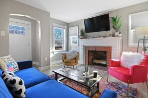 A living room with a blue sofa, a red armchair, a fireplace, and a television mounted above it. - Walk Downtown From Wonderland! (Columbia)