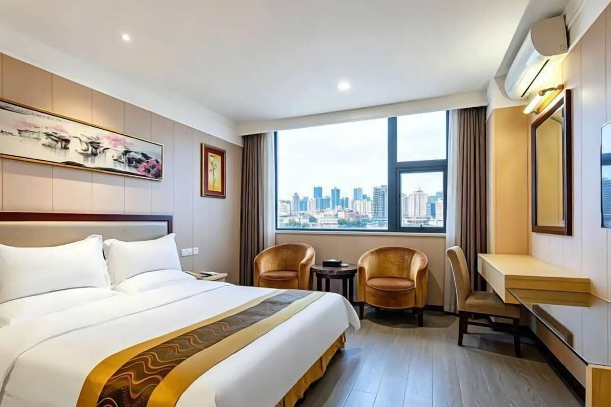 A hotel room with a large bed, two armchairs, a small table, a desk, a large window with a city view, and artwork on the walls.