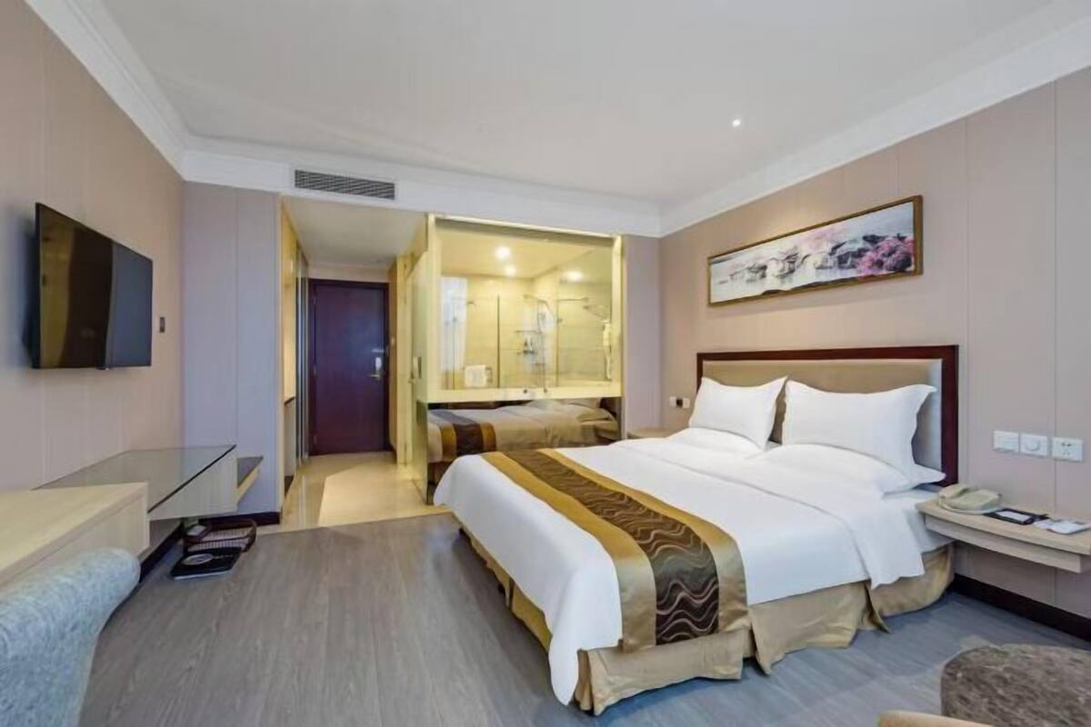 A hotel room with a bed, bedside table, TV, and a bathroom with a glass shower enclosure.