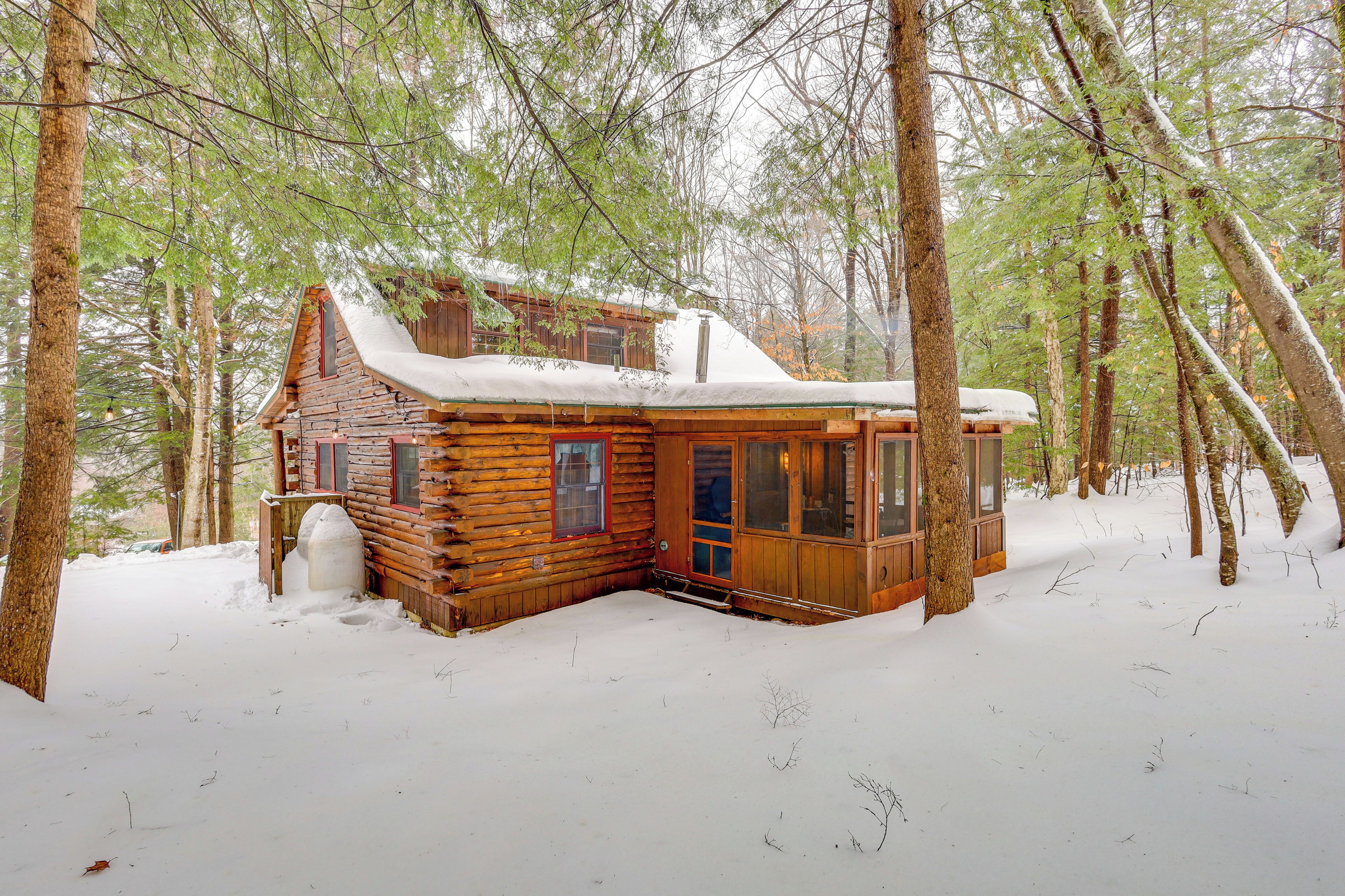 Uma cabana de tronco com telhado coberto de neve, rodeada por árvores altas numa floresta.