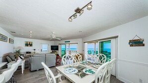 Uma área de refeições com uma mesa posta para quatro pessoas, rodeada por cadeiras brancas com assentos trançados.