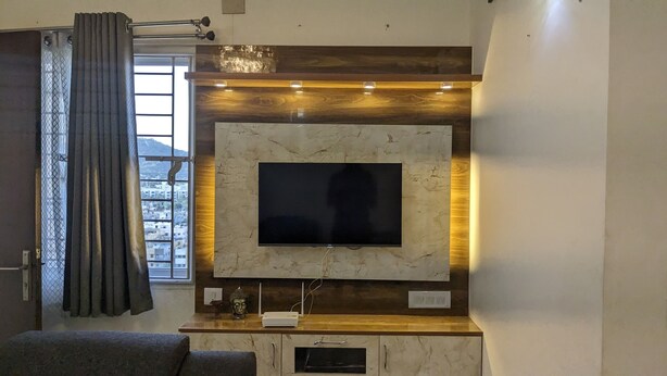 A modern living room with a flat-screen TV mounted on a marble wall, a wooden entertainment unit below, and a large window with curtains. - Bed and Brew (Chennai)