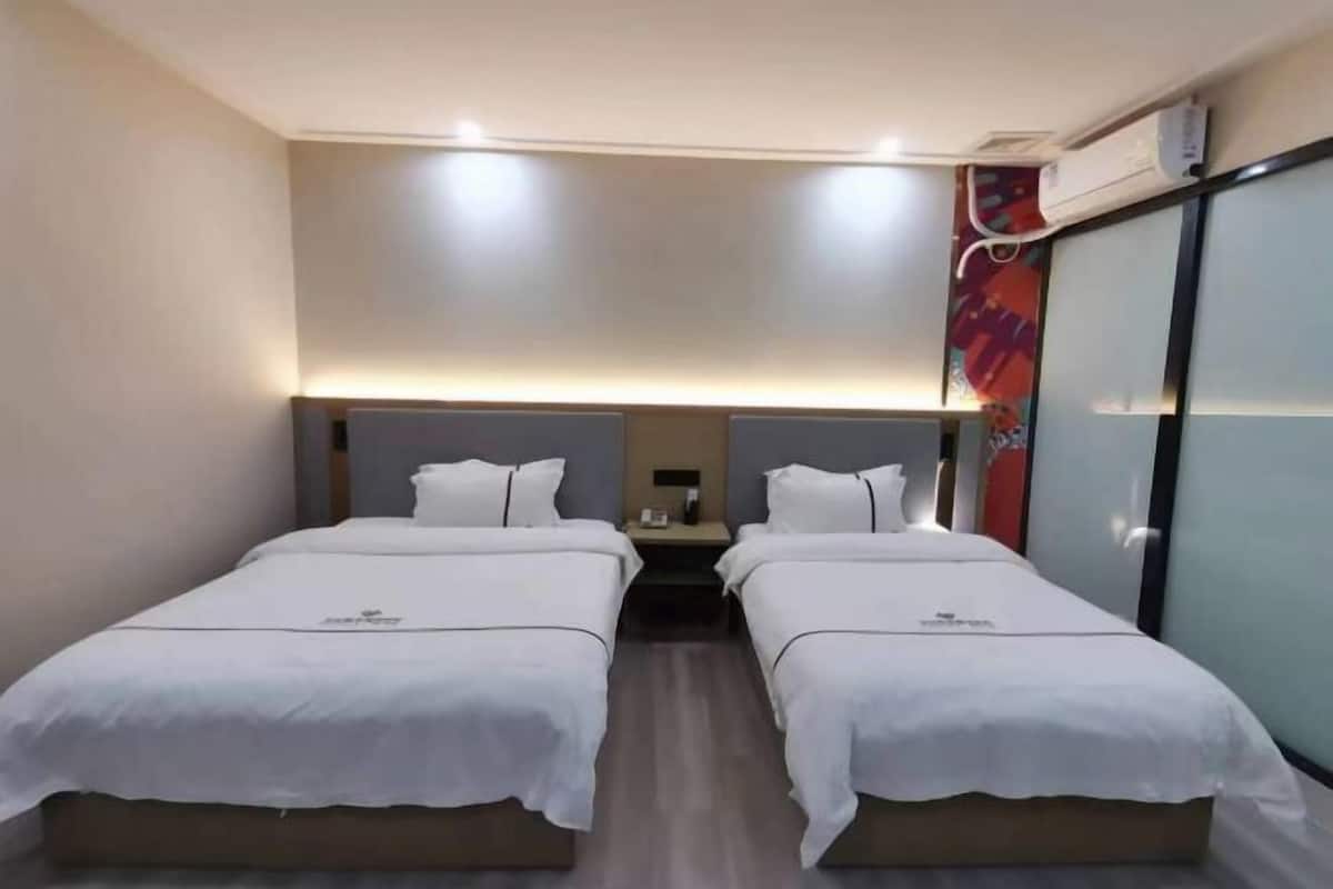 A hotel room with two beds, a nightstand, a wall-mounted air conditioner, and a glass partition.
