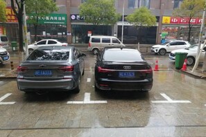 A parking lot with cars parked in designated spaces, a wet surface indicating recent rain, and a building with signage in a foreign language in the background. - Century Business Hotel (Zuoling New Town) (Wuhan)