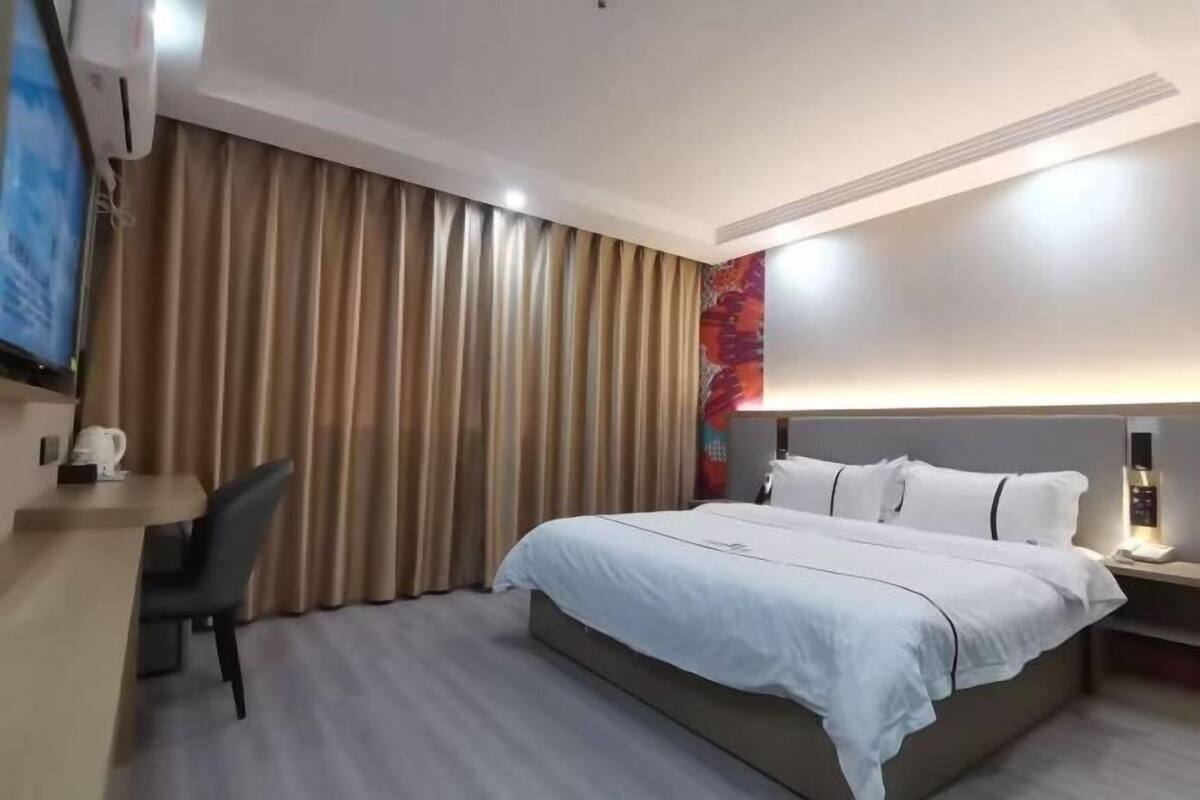 A hotel room with a bed, a desk, a chair, curtains, a wall-mounted TV, and a bedside table with a lamp.