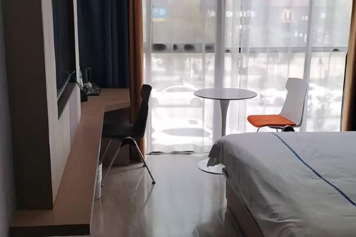 A hotel room with a bed, a desk, a chair, a small table, and a view of the street through large windows.