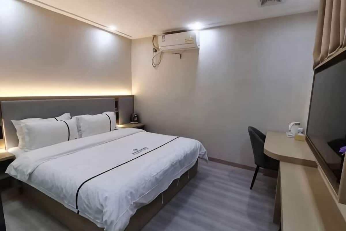 A hotel room with a bed, bedside table, desk, chair, and an air conditioning unit.