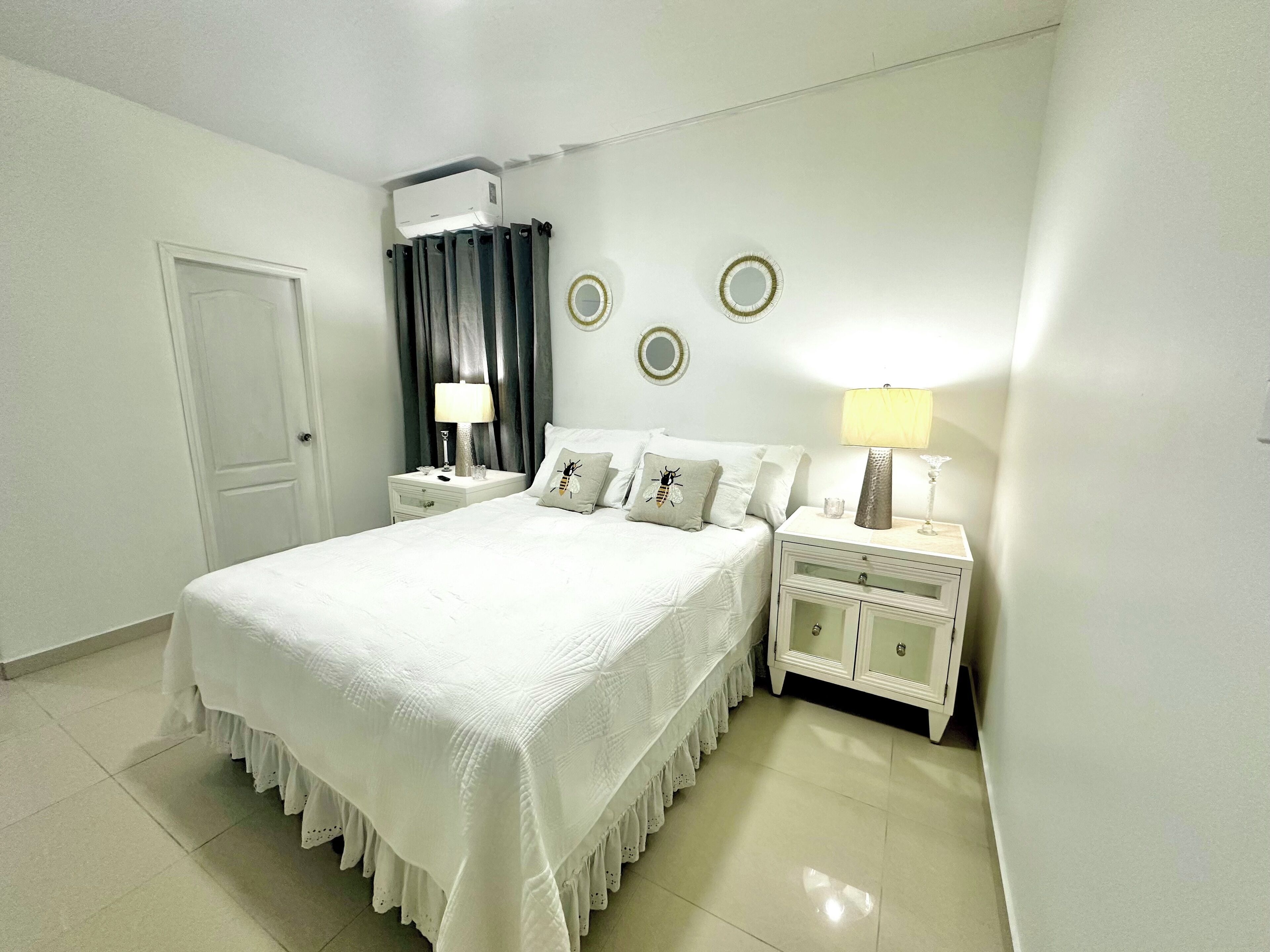 A neatly arranged bedroom with a large bed, two bedside tables, a nightstand, a door, and a wall-mounted air conditioner.
