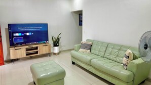 Ein Wohnzimmer mit einem grünen Sofa, einem passenden Hocker, einem Flachbildfernseher auf einem Holzboden, einer Topfpflanze und einem Standventilator.