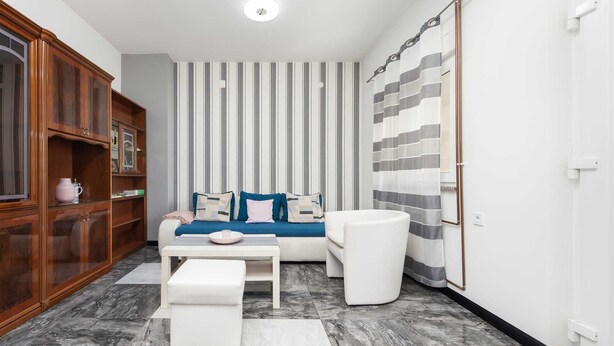 Un salon moderne avec un canapé, un pouf et une table basse. On y trouve un rideau à rayures et un meuble en bois doté de portes en verre.