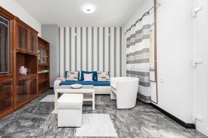 Un salon moderne avec un canapé, un pouf et une table basse. On y trouve un rideau à rayures et un meuble en bois doté de portes en verre.