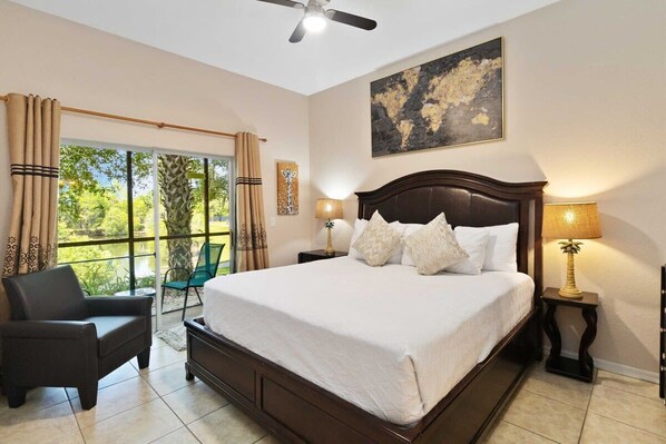A bedroom with a large bed, a chair, a ceiling fan, and a view of greenery through a window.