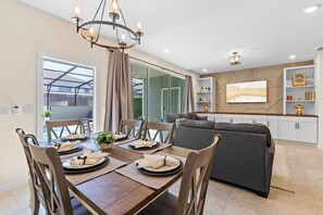 A dining area with a wooden table, chairs, and a chandelier. There is a sofa and a TV in the background. - Amazing 5BR house with theme rooms, POOL HEAT INCLUDED (Davenport)