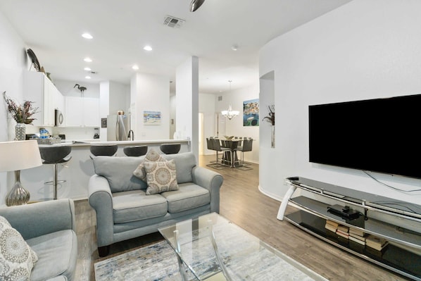 A modern living room with a grey sofa, a glass coffee table, and a flat-screen TV mounted on a wall.