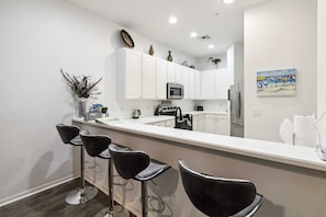 A modern kitchen with white cabinets, a microwave, and a refrigerator. There are three black bar stools and a vase with dried plants on the counter.