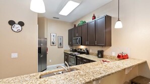 Une cuisine dotée de plans de travail en granit, de meubles de couleur brun foncé, d’un réfrigérateur en acier inoxydable, d’un four à micro-ondes, d’une machine à café et d’une horloge décorée avec un motif de Mickey Mouse.