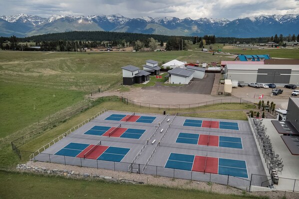 An outdoor tennis court with a mountainous backdrop, surrounded by buildings and a parking area.