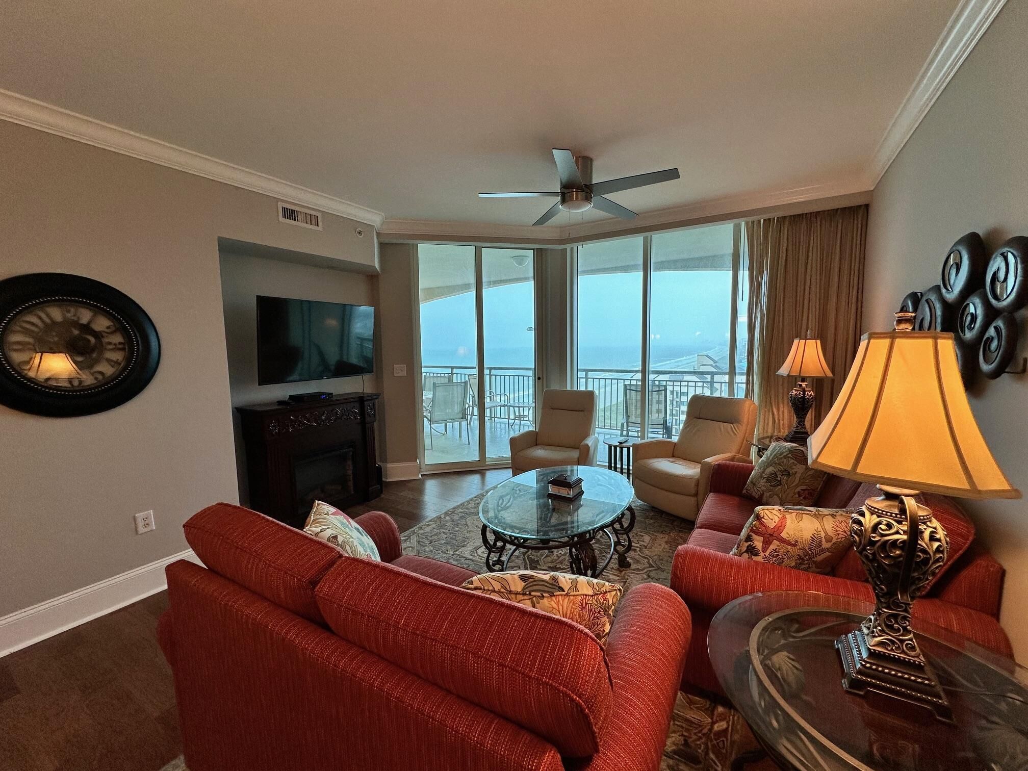 A spacious living room with a flat-screen TV, a fireplace, and a large window offering a view of the ocean. There are two sofas, a coffee table, and a lamp.